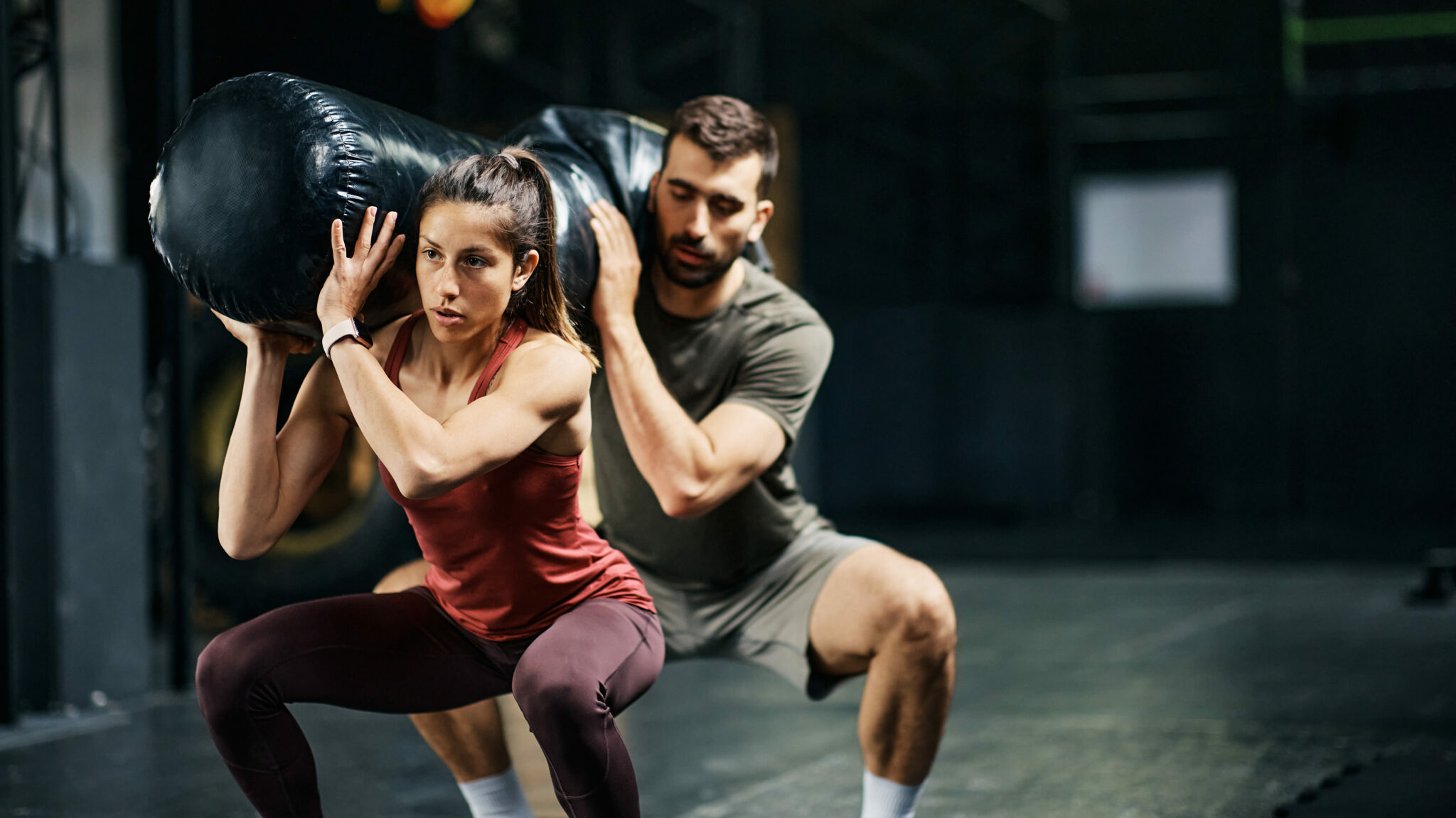 Athletic couple doing squats while holding sandbag on shoulders during gym workout. Focus is on woman.